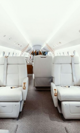 Interior of a private jet featuring large white leather seats arranged facing each other, a central aisle, and windows on both sides, creating a bright and luxurious atmosphere.