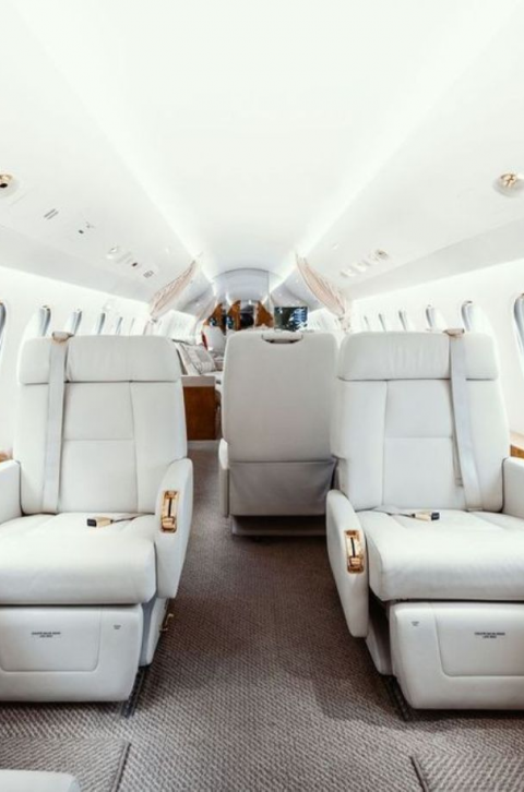 Intérieur d’un jet privé avec de larges sièges en cuir blanc disposés face à face, un couloir central et des hublots alignés de chaque côté, créant une ambiance lumineuse et luxueuse.