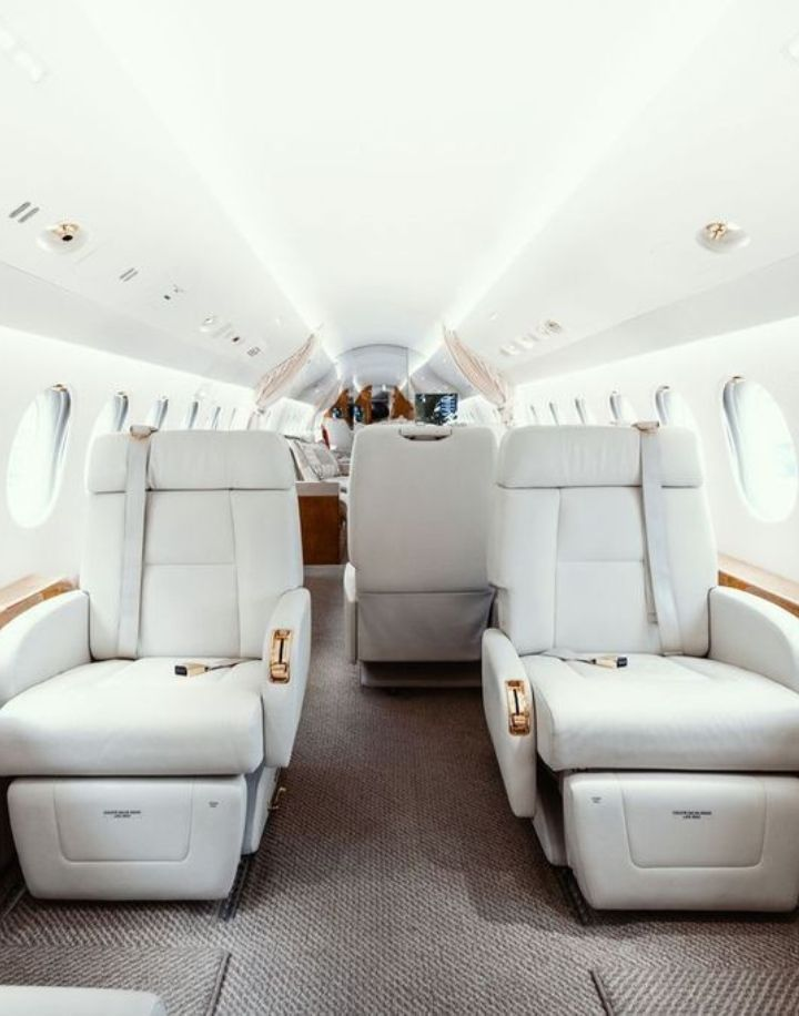 Interior of a private jet featuring large white leather seats arranged facing each other, a central aisle, and windows on both sides, creating a bright and luxurious atmosphere.