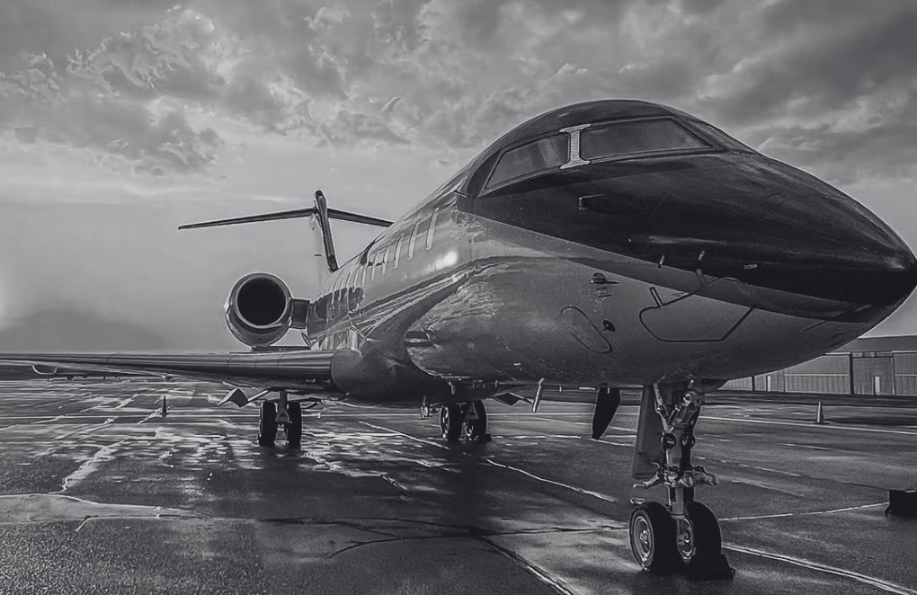 Photo en noir et blanc d'un jet privé d'affaires stationné sur le tarmac d'un aéroport, avec son train d'atterrissage déployé et un ciel nuageux dramatique en arrière-plan.