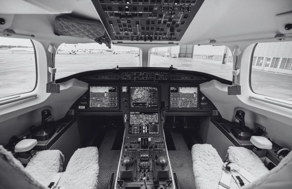 Modern aircraft cockpit with multiple navigation screens, central console equipped with flight controls, pilot seats covered with white sheepskin covers, and large windows offering a view of the tarmac.